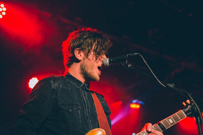 Man on stage singing into a mike with red lights in background